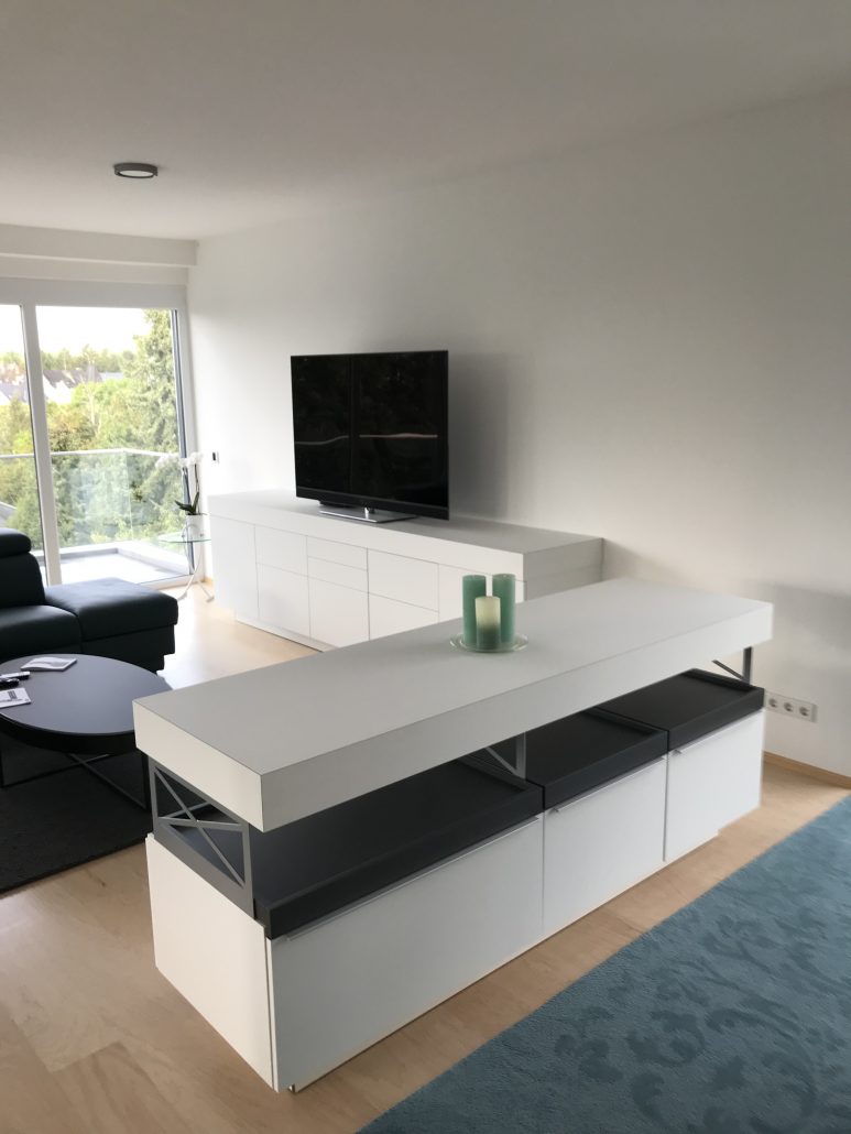 Ein weißes Sideboard mit dunklen Einschüben, dahinter ein weiteres weißes Sideboard mit einem Fernseher darauf.